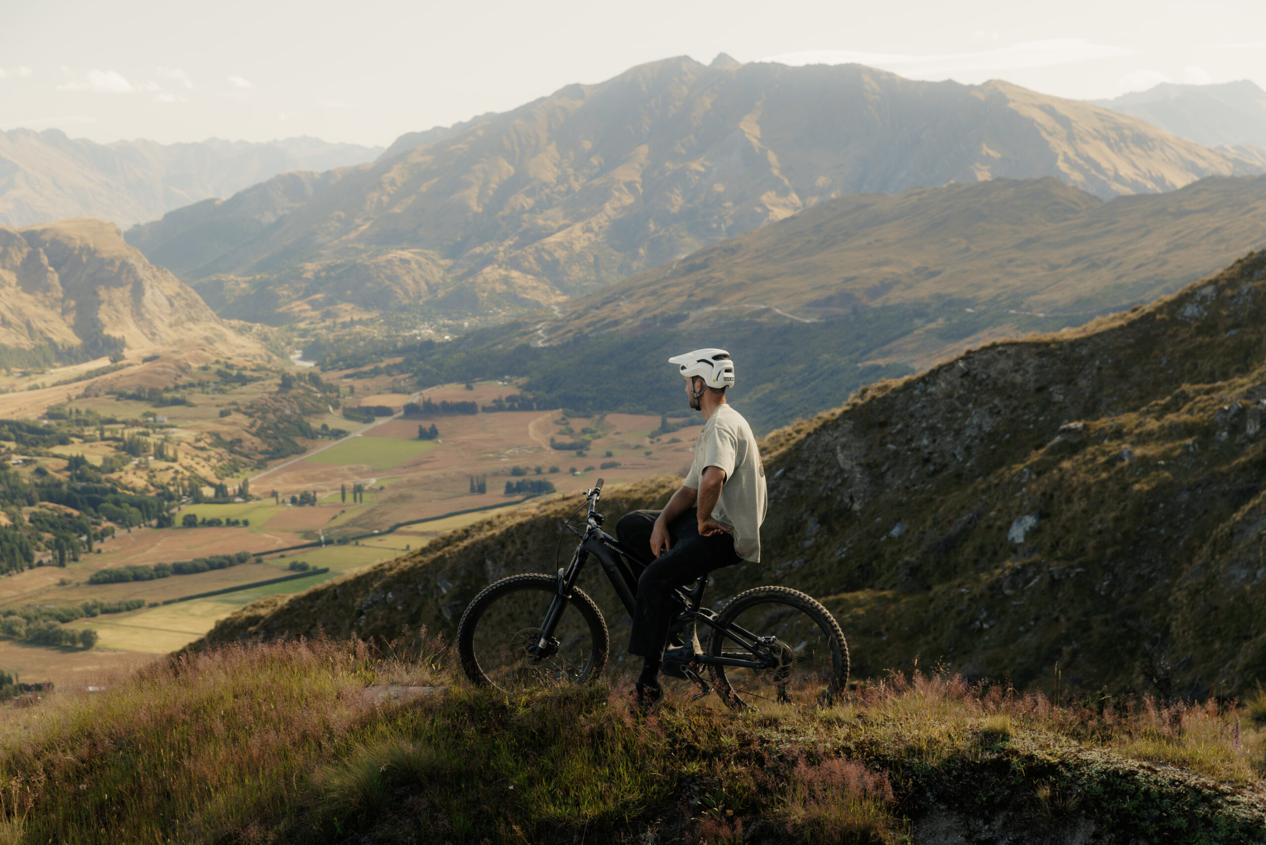 Man wearing a white helmet standing beside a mountain bike on a hillside, with a valley and distant mountains in the background