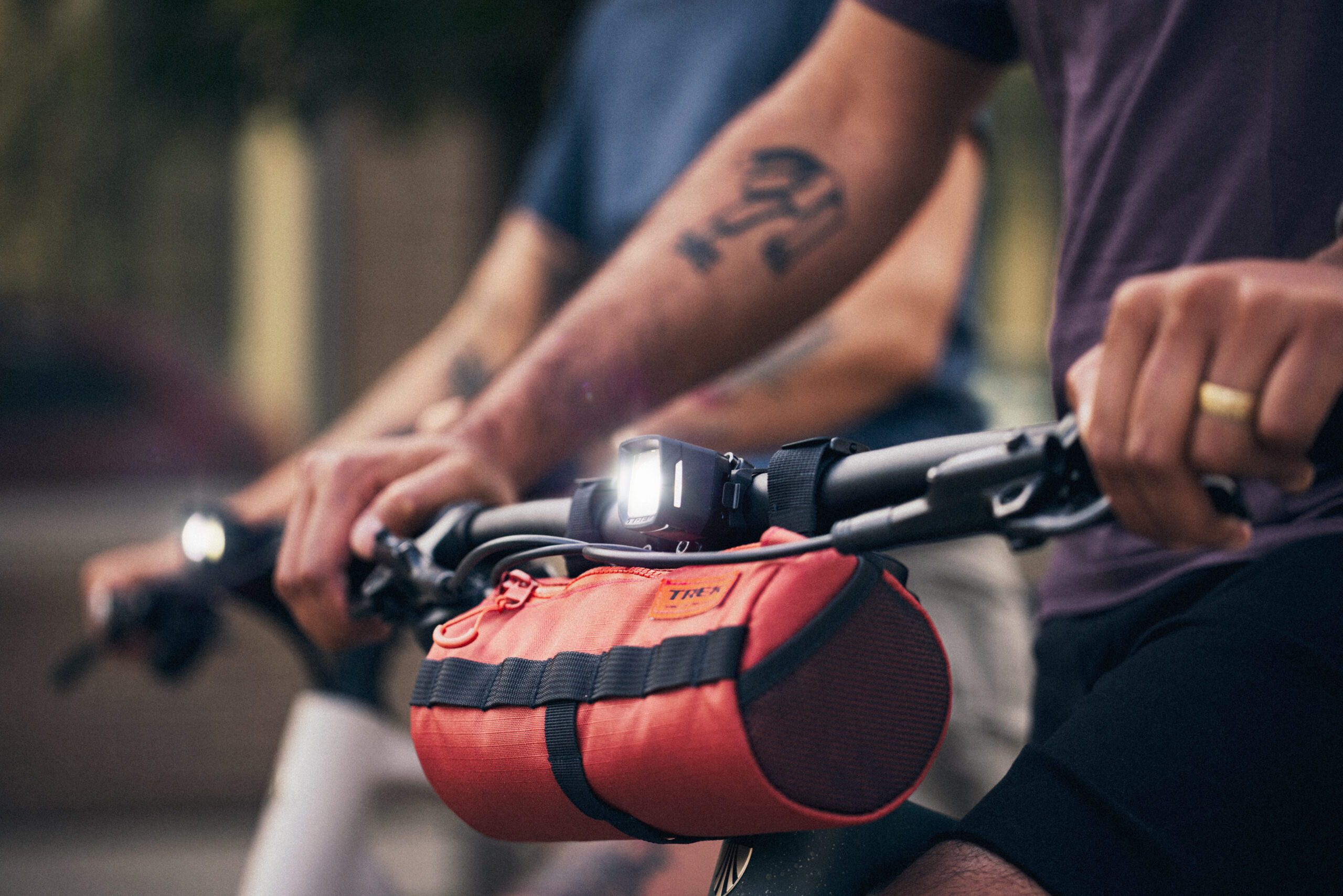 People riding bikes, close-up of arms on handlebars with a red vest/bag in the foreground and tattoos on the forearm visible behind.