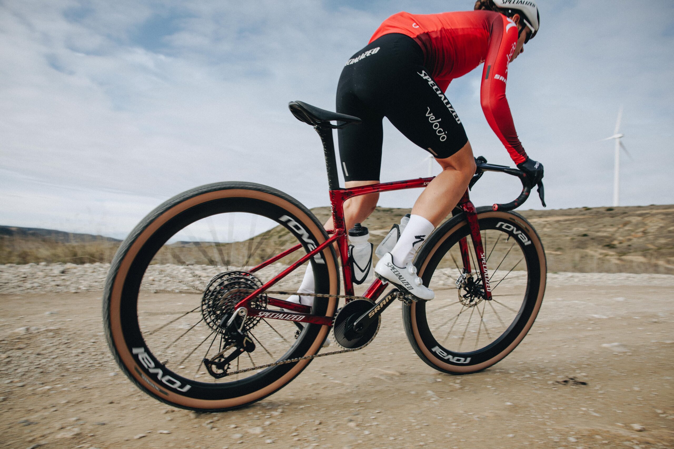 Cyclist in red jersey riding a red road bike on a dry rural road with clear skies and a distant wind turbine on the horizon.