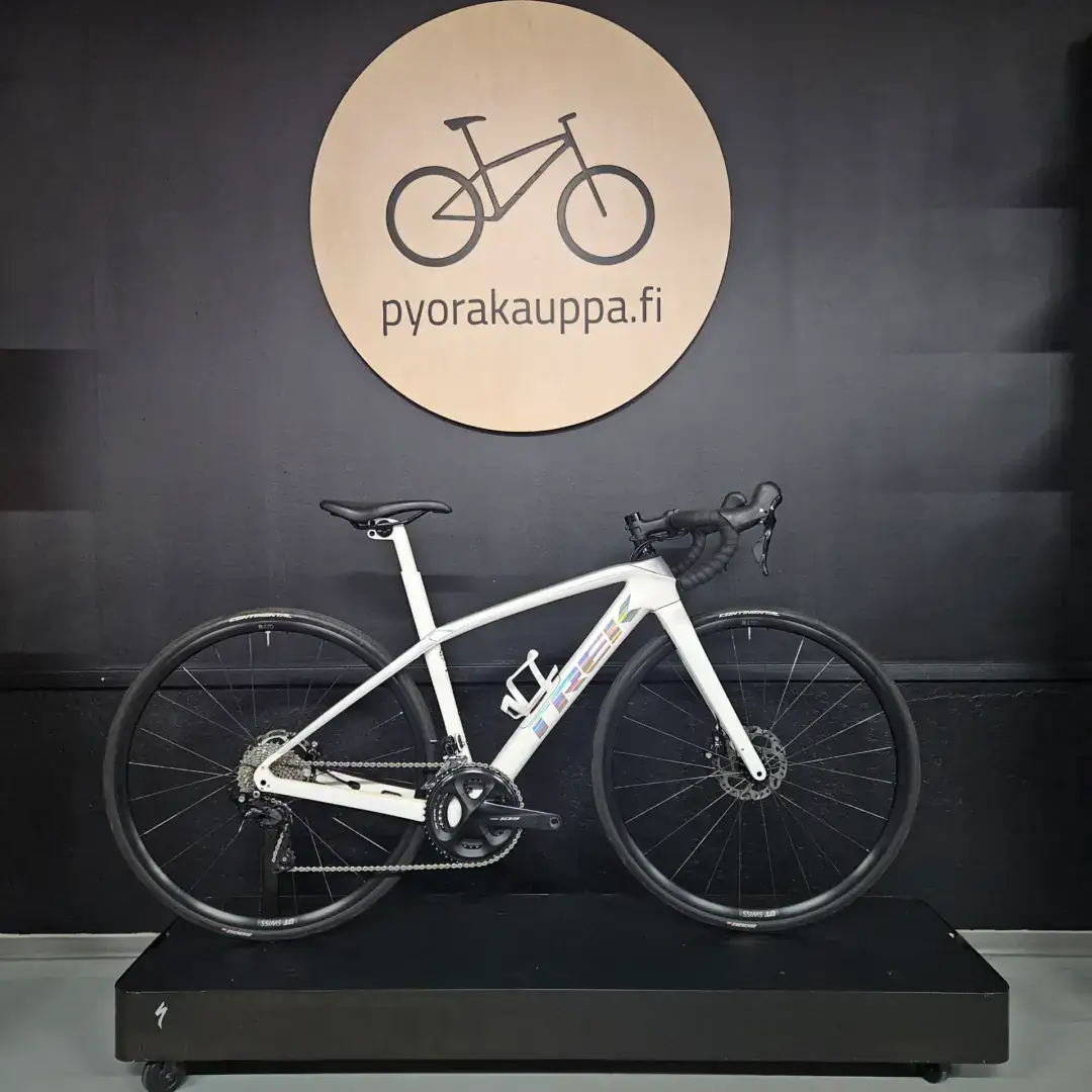 White road bicycle mounted on a black display platform in front of a dark wall, with a round wooden sign reading 'pyorakauppa.fi' behind it.