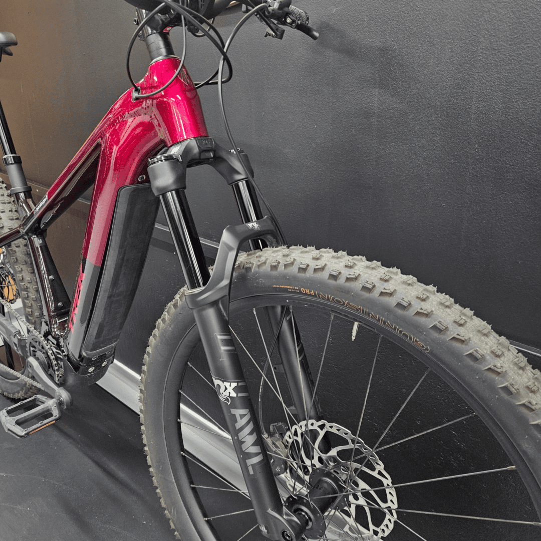 Close-up of a red mountain bike frame with front suspension fork, hydraulic disc brakes, and knobby tires against a dark wall surface.