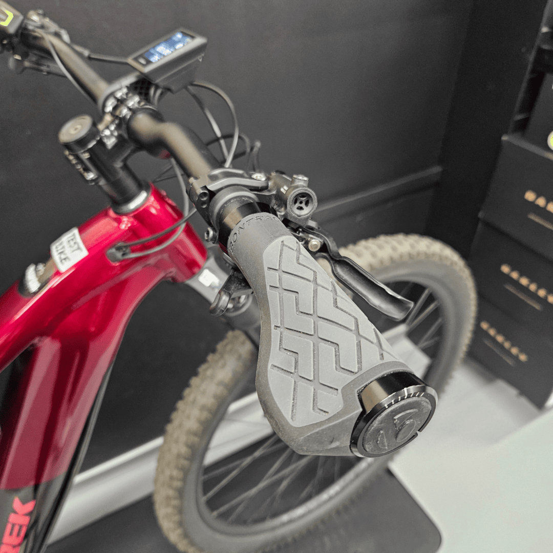 Close-up of a bike handlebar area showing a mounted bike computer, cables, and red frame with a front tire in view.