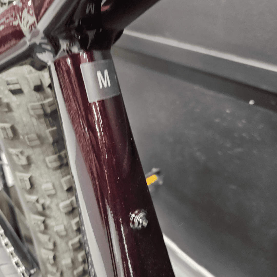 Close-up of a maroon bicycle frame with a size tag reading 'M' attached near the seat tube or down tube.