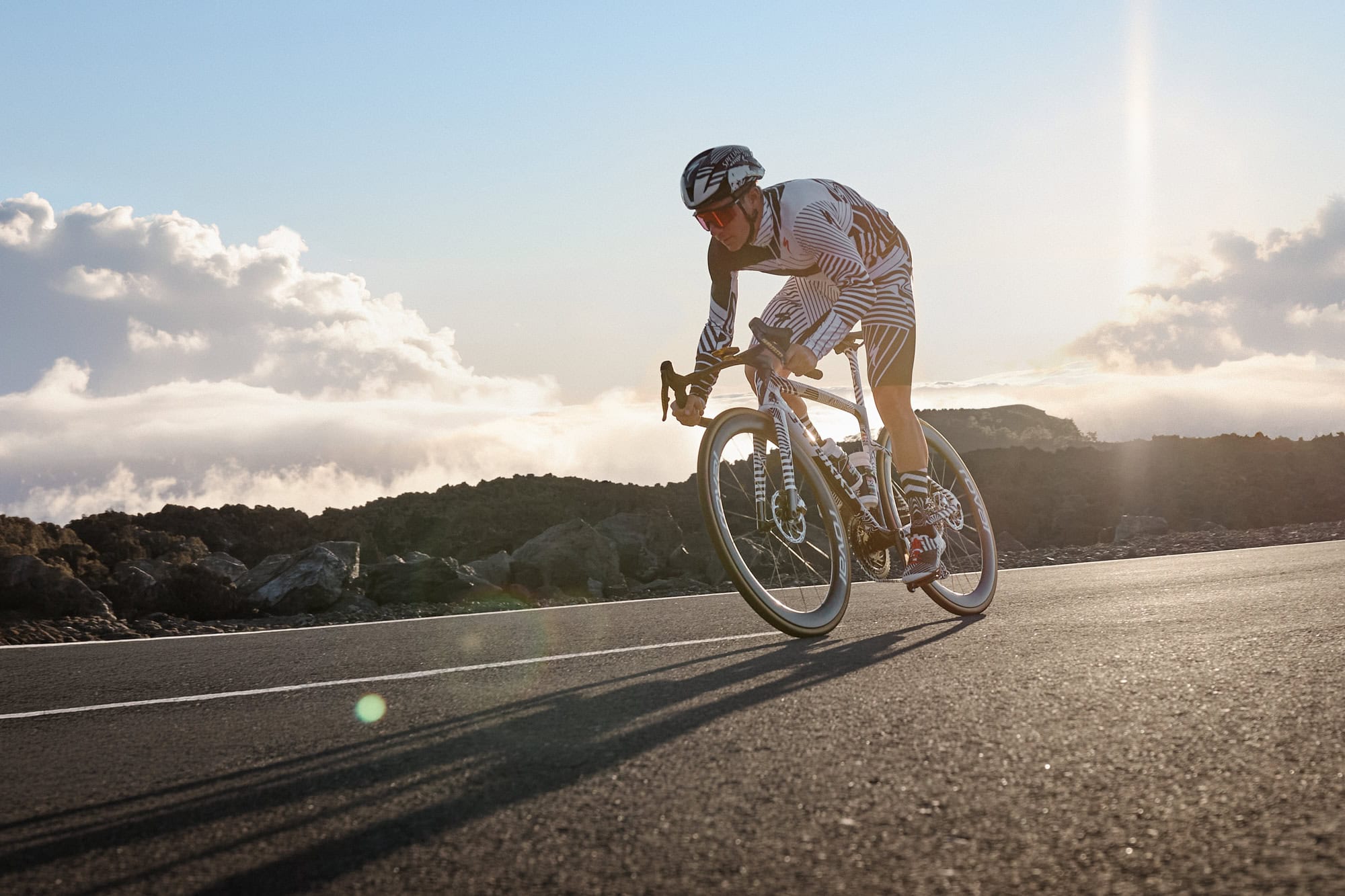Cyclist in matching black-and-white kit riding a racing bike on a coastal road at sunset, rocks in the foreground and clouds in the sky behind.