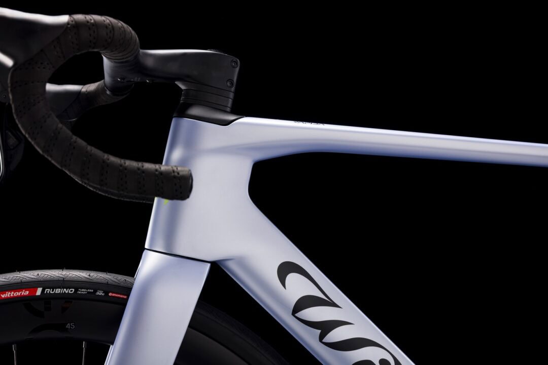 Close-up of a road bike cockpit: white aero frame, compact handlebars with perforated tape, and a tubeless tire against a black backdrop, highlighting the modern design.