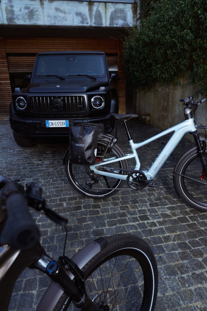 Black Mercedes G‑Wagon parked in a cobblestone driveway beside a blue electric bike with a rear pannier bag in the foreground.