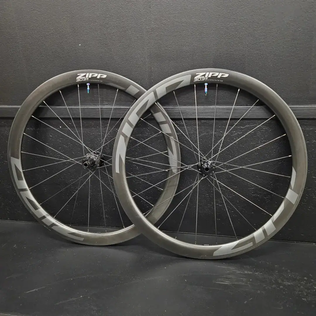 Two carbon bike wheels with deep rims leaning against a dark wall, showing Zipp branding on the rims.