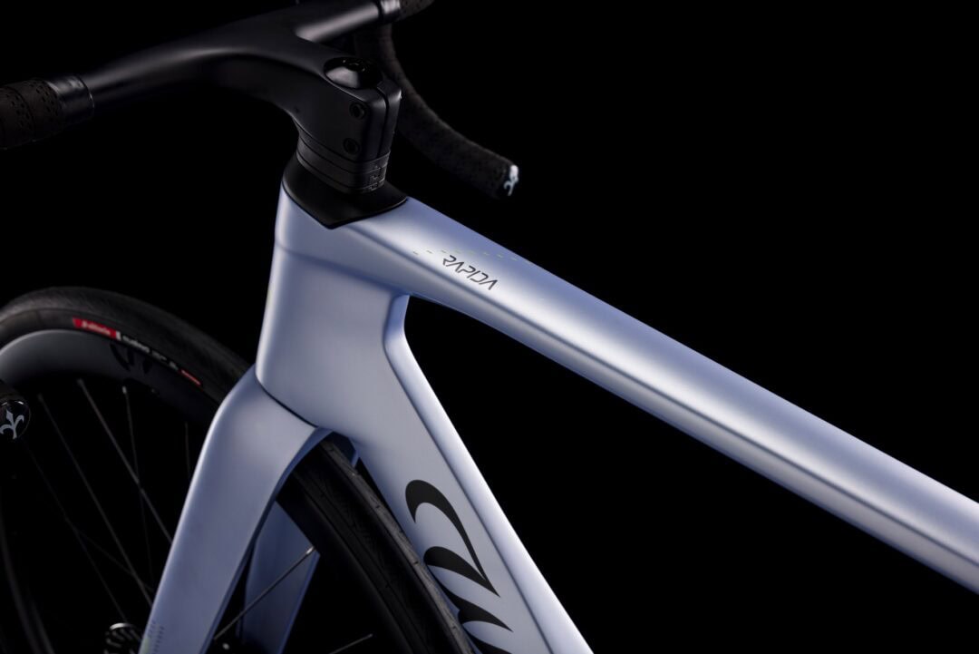 Close-up of a pale blue road bike frame near the headset and handlebars, with a front wheel in view against a black background.