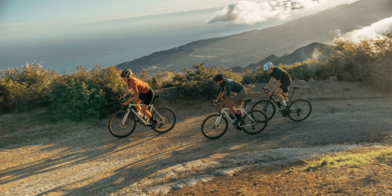 Three mountain bikers wearing helmets ride along a sunlit dirt trail with coastal mountains in the background.