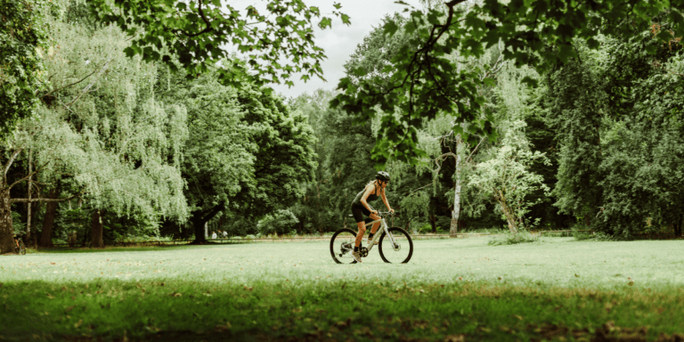 Cyclist wearing a helmet rides a bike across a grassy park with trees surrounding the area.