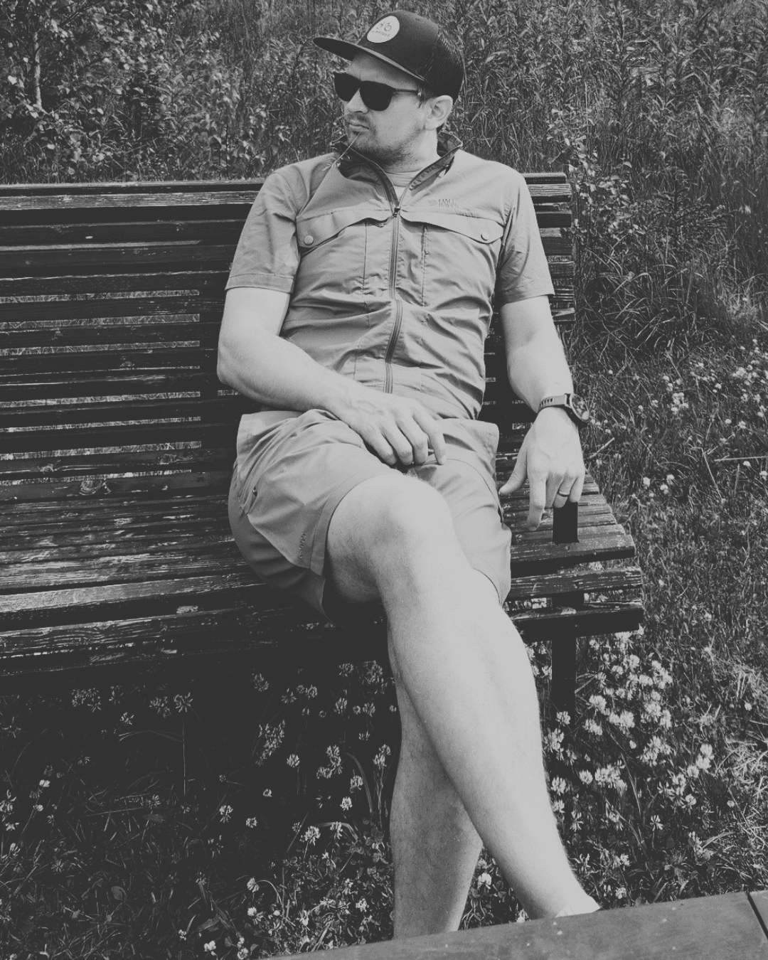 Man in a cap and sunglasses sits relaxed on a wooden park bench, leaning back outdoors with tall grass in the background.