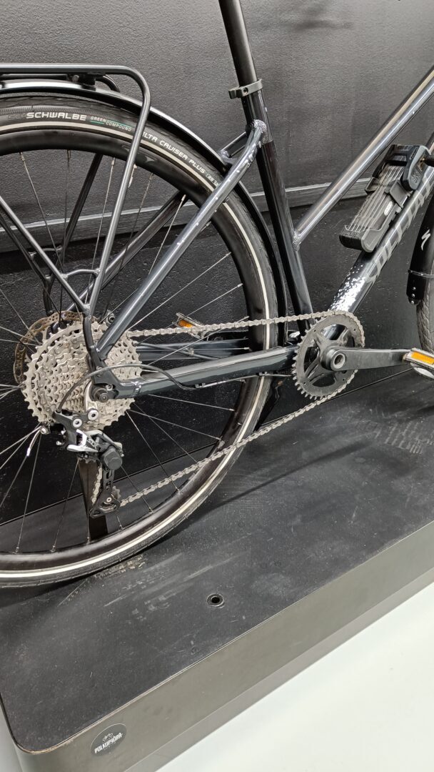 Rear-side view of a gray bicycle showing the cassette, chain, derailleur, and part of the frame with a mounted rack and battery pack on an indoor display wall.