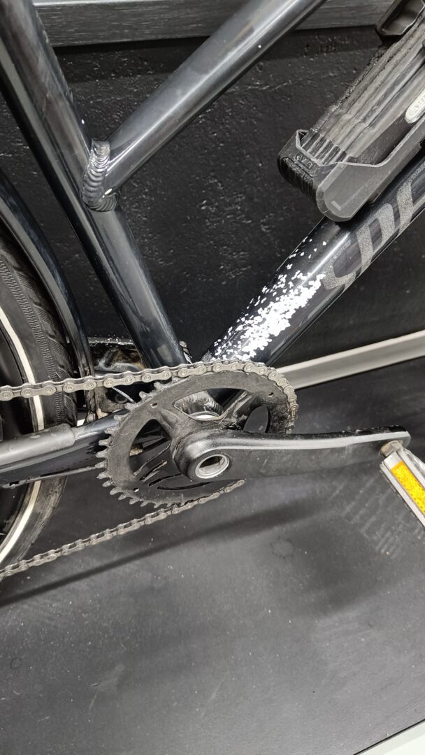 Close-up of a bicycle crankset and chain on a dark frame, with a worn pedal and frame scratches visible at the bottom bracket area