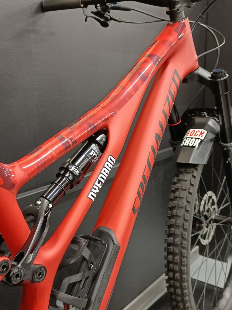 Close-up of a red full-suspension mountain bike frame, showing the top and down tubes and a RockShox front fork with a knobby tire nearby
