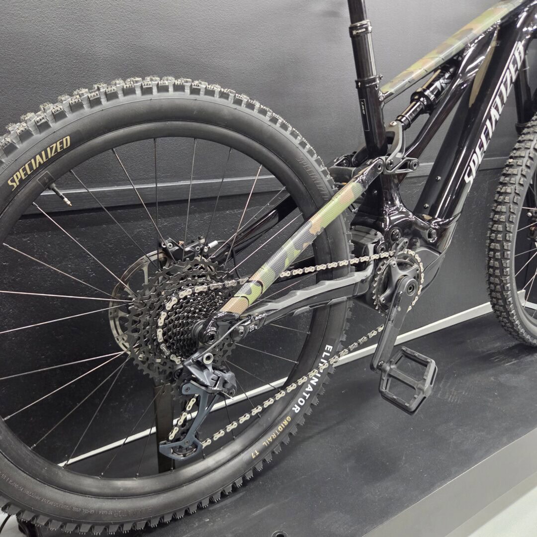 Close-up of a mountain bike's rear wheel and drivetrain, showing a knobby tire, cassette, derailleur, and part of a camouflaged frame.