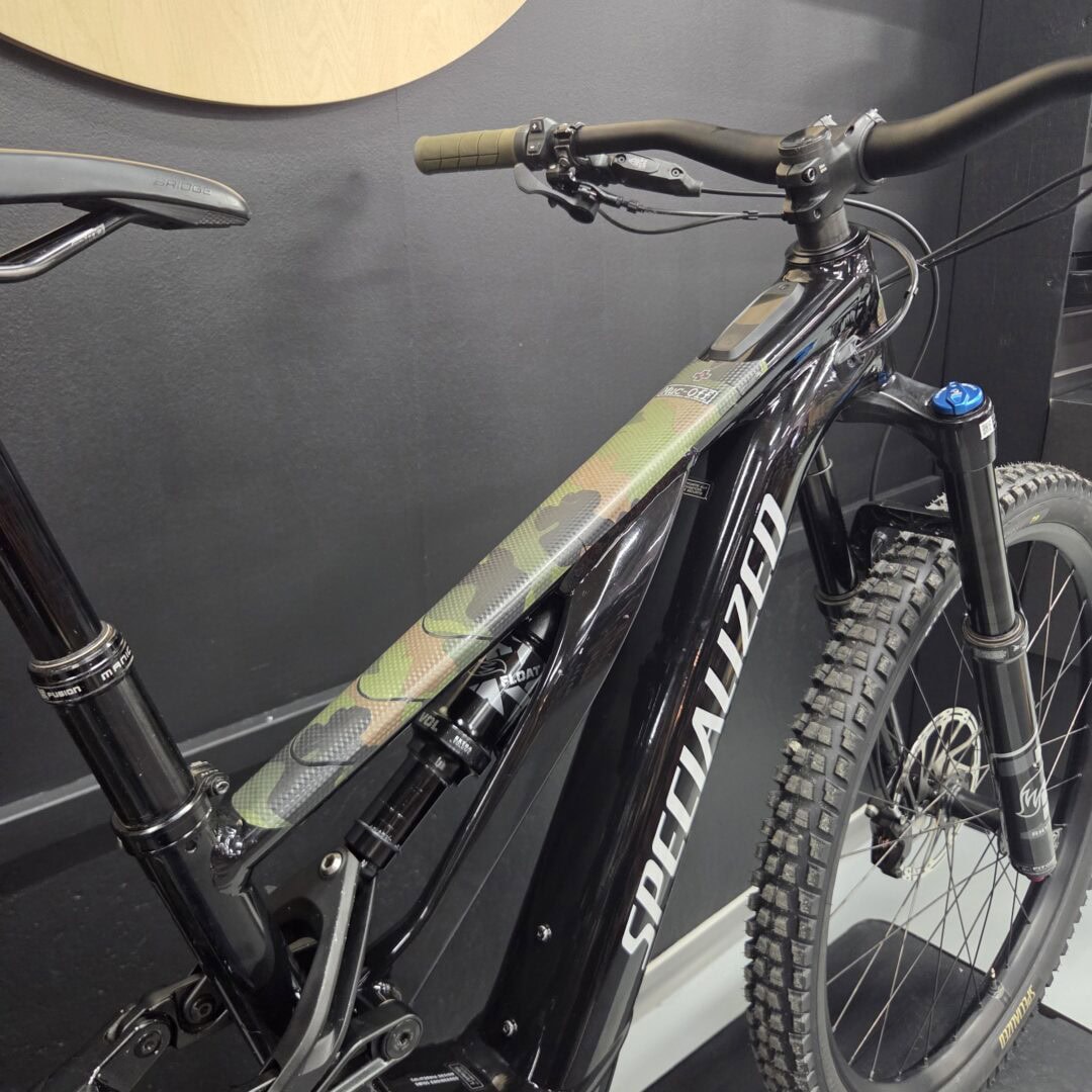 Close-up of a black mountain bike frame with green camouflage stripe, front suspension fork, handlebar and knobby tire visible.