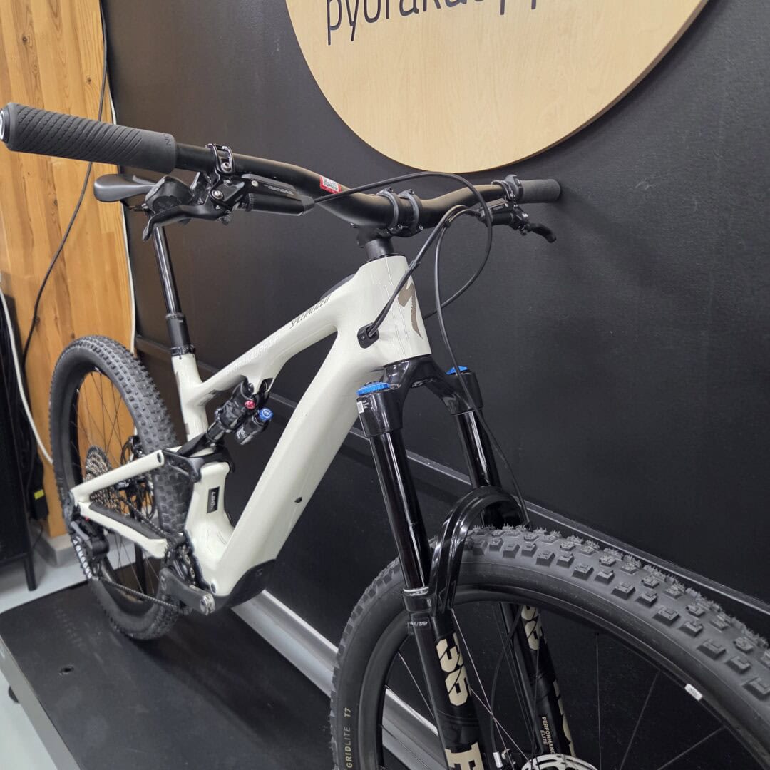 Grey full-suspension mountain bike indoors, showing wide knobby tires, front suspension fork, and handlebar controls.