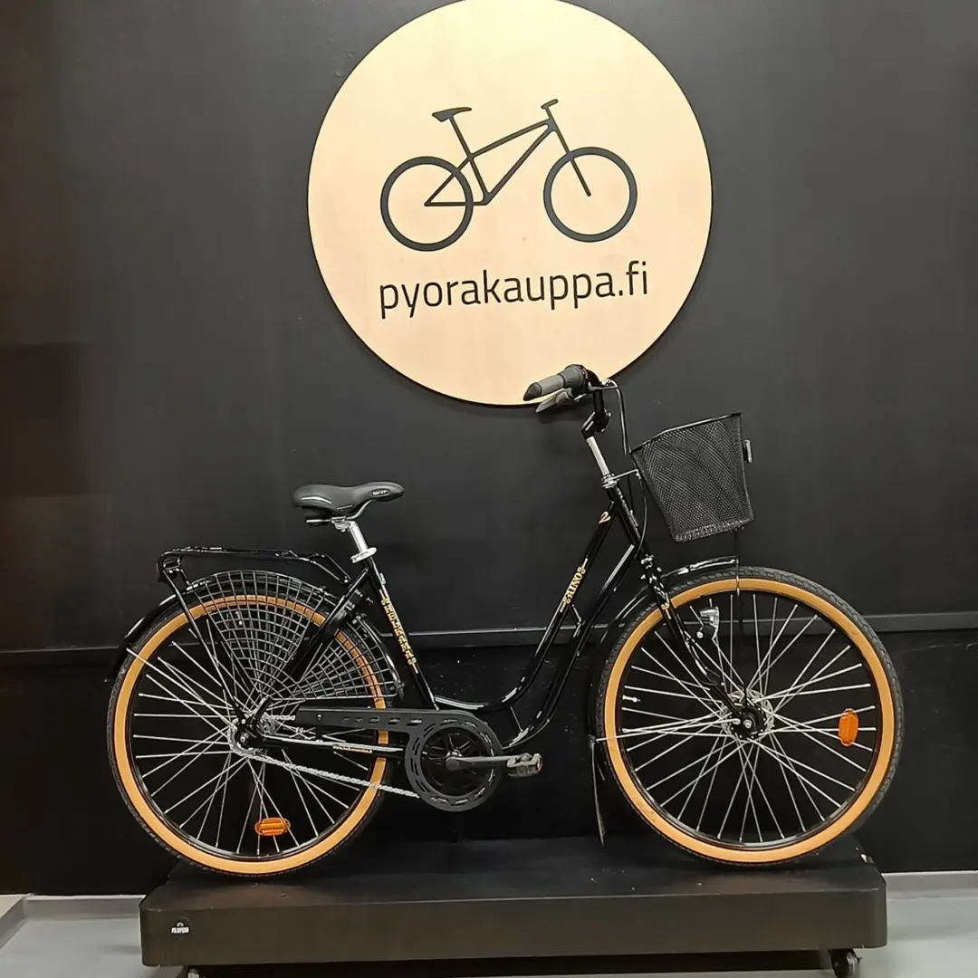 Two black bicycles with tan tires displayed on a platform against a dark wall, a round wooden sign behind them reading 'pyorakauppa.fi'.