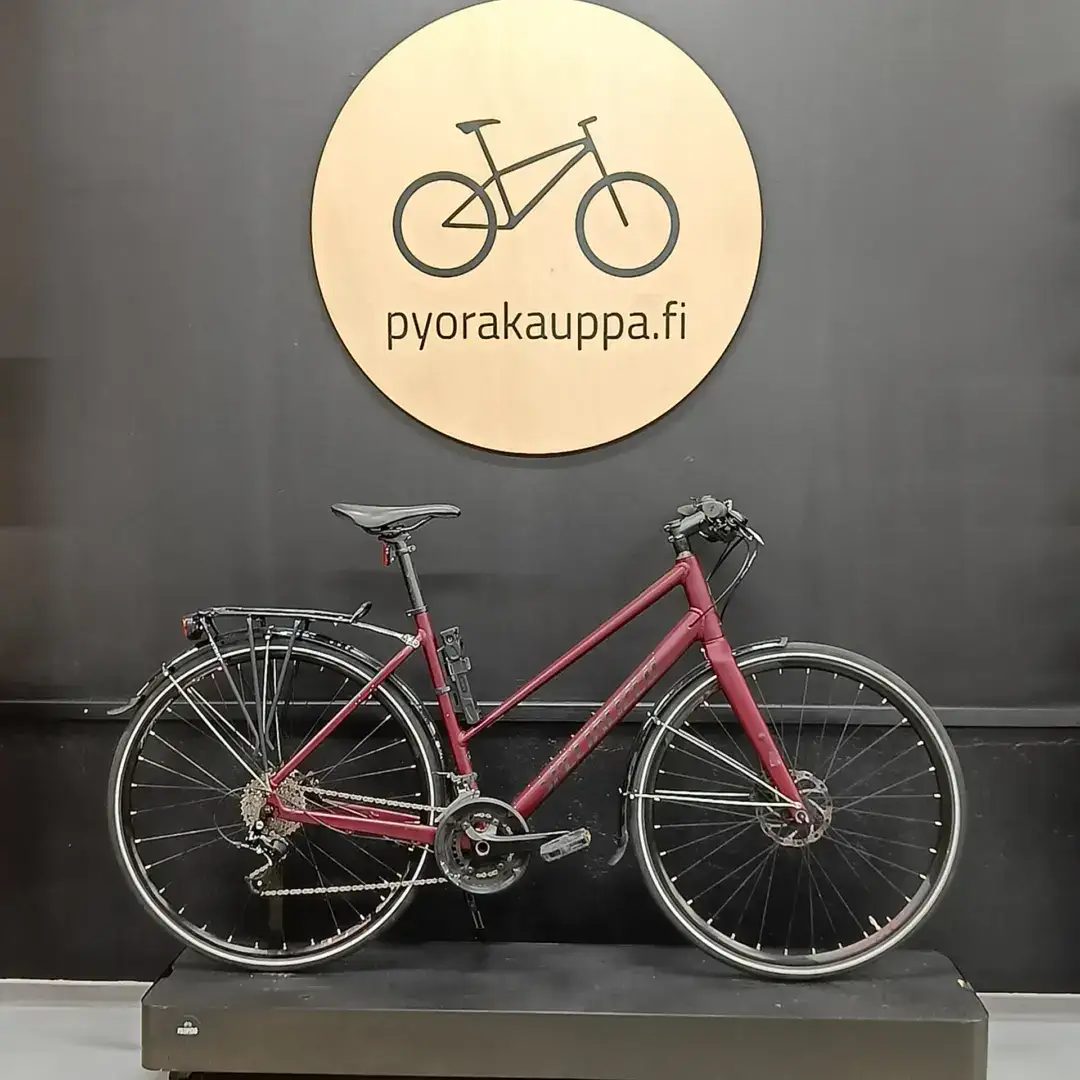 Red bicycle on a black display stand in front of a large circular wooden sign that reads 'pyorakauppa.fi'
