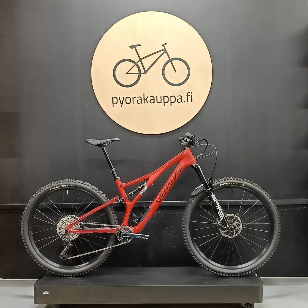 Red full-suspension mountain bike on a display stand against a dark wall, with a round light wood sign reading 'pyorakauppa.fi' above it.