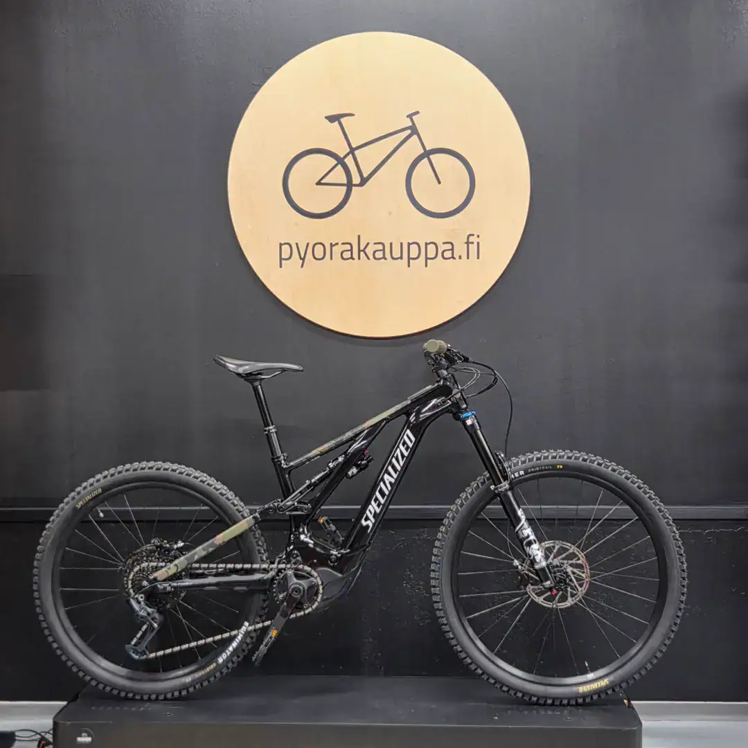 Black full-suspension mountain bike on display against a dark wall, with a round wooden sign reading 'pyorakauppa.fi' behind it.