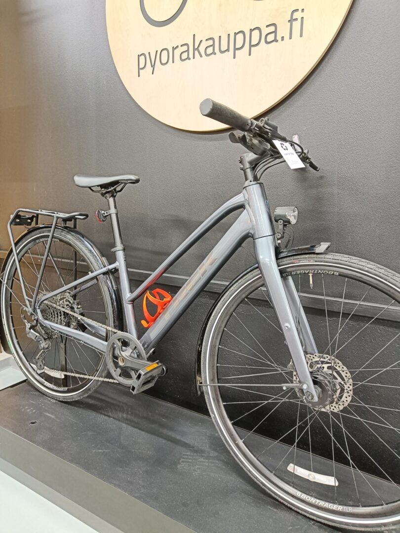 Grey hybrid bicycle on display indoors with a round wooden sign behind reading pyorakauppa.fi