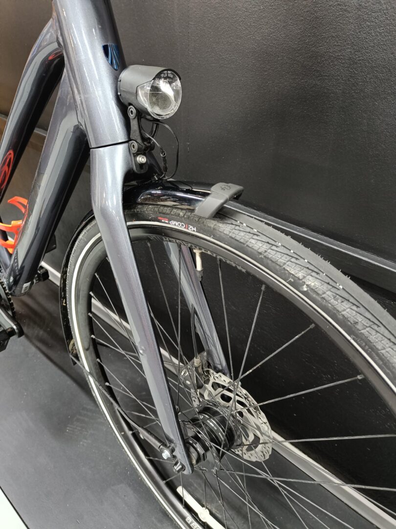 Front wheel and fork of a bicycle with a mounted headlight above the front fender and visible brake rotor, cables, and tire detailing along a dark wall.