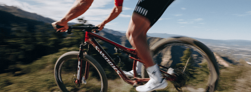 Action shot of a cyclist riding a red mountain bike on a dirt trail with mountains in the background, conveying speed and motion.