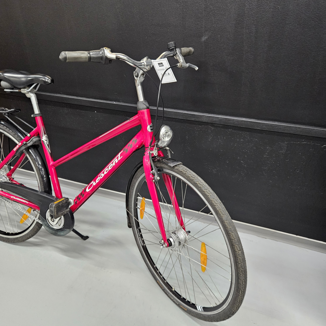 Pink city bike with a black saddle and chrome handlebars, leaning against a dark wall; front wheel visible in the foreground.