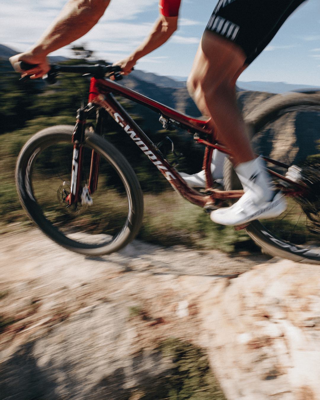 Mountain biker riding a red bike down a rocky trail, legs blurred in motion against a mountainous landscape.