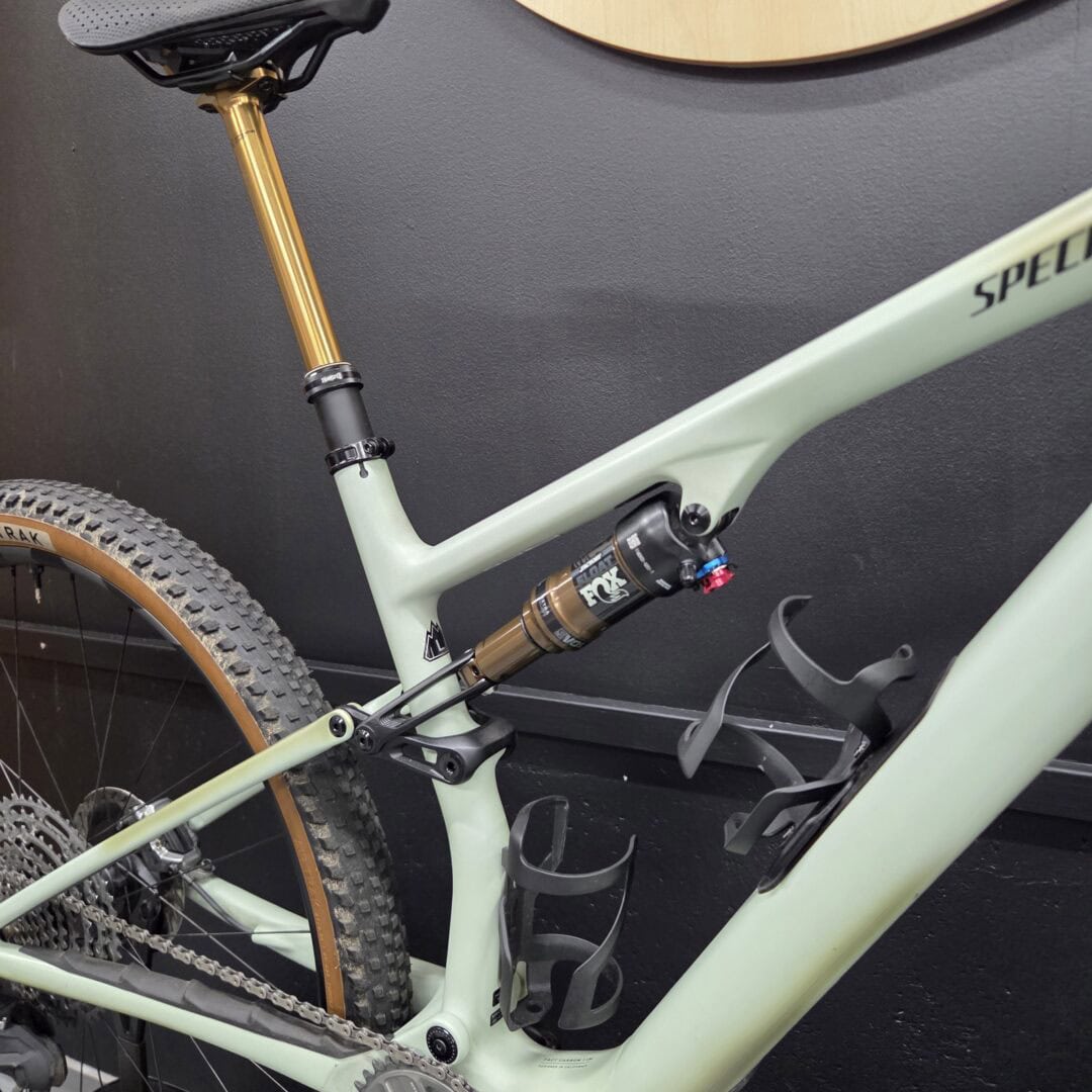 Close-up of a mint-green mountain bike frame with a gold suspension fork, rear shock, and chain near the drivetrain against a dark wall.