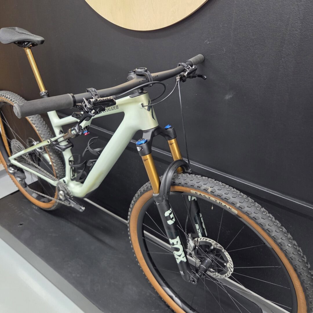 Close-up of a mint-green Specialized mountain bike showing handlebars, front suspension fork, and knobby tires against a dark wall.