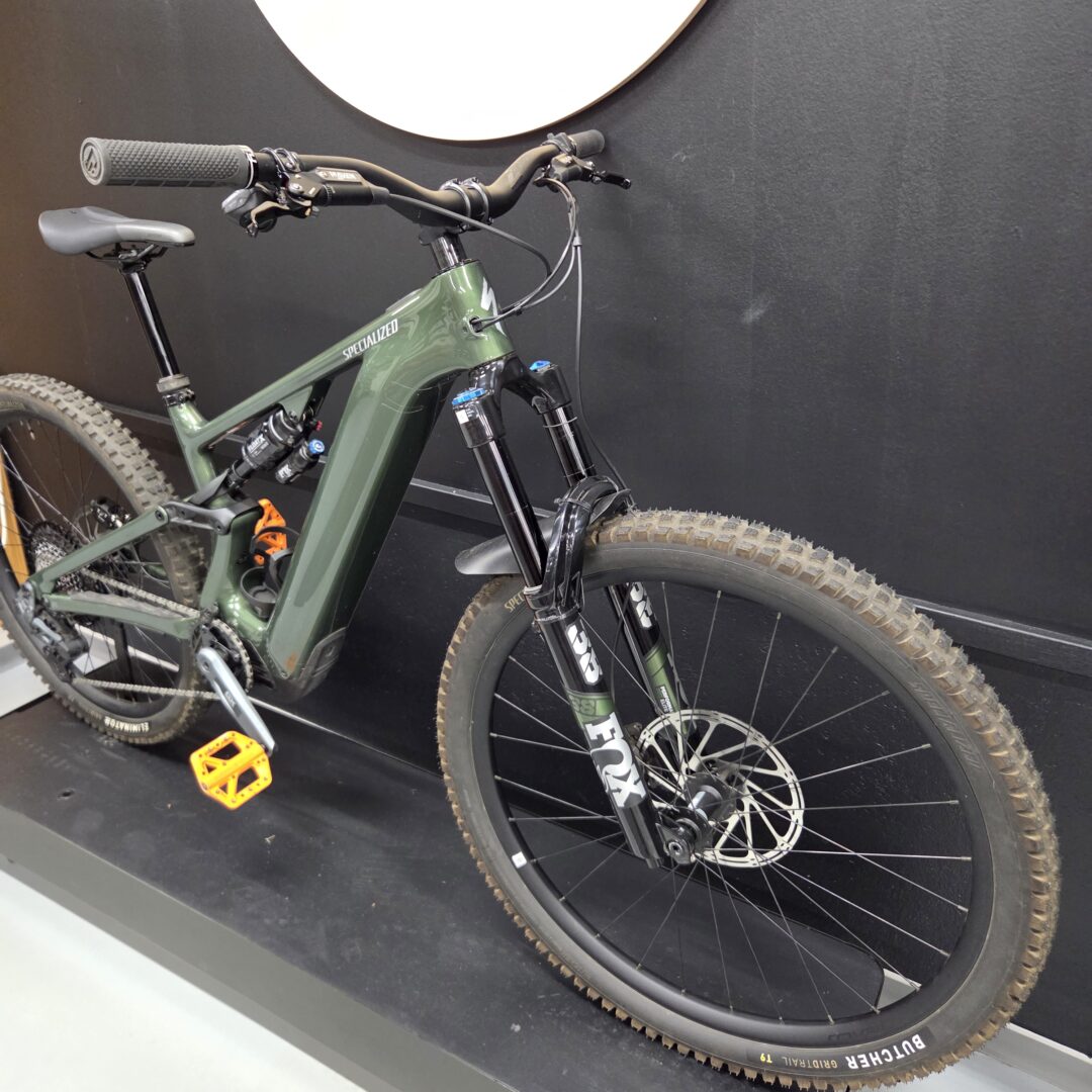 Green Specialized mountain bike with front suspension, parked indoors against a dark wall; orange pedals and knobby tires visible.