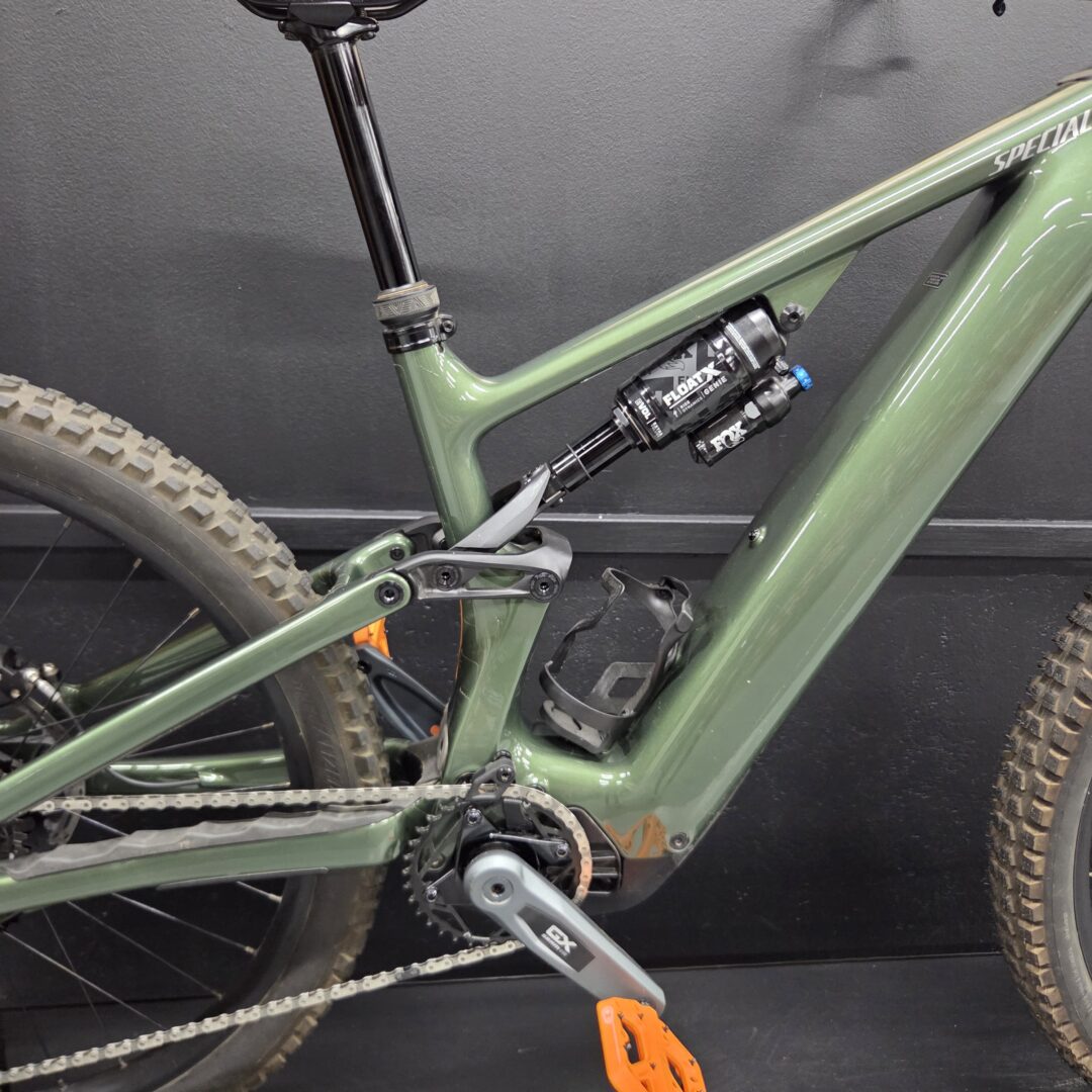 Close-up of a green full-suspension mountain bike frame with an orange pedal, drivetrain, and rear shock against a dark background.