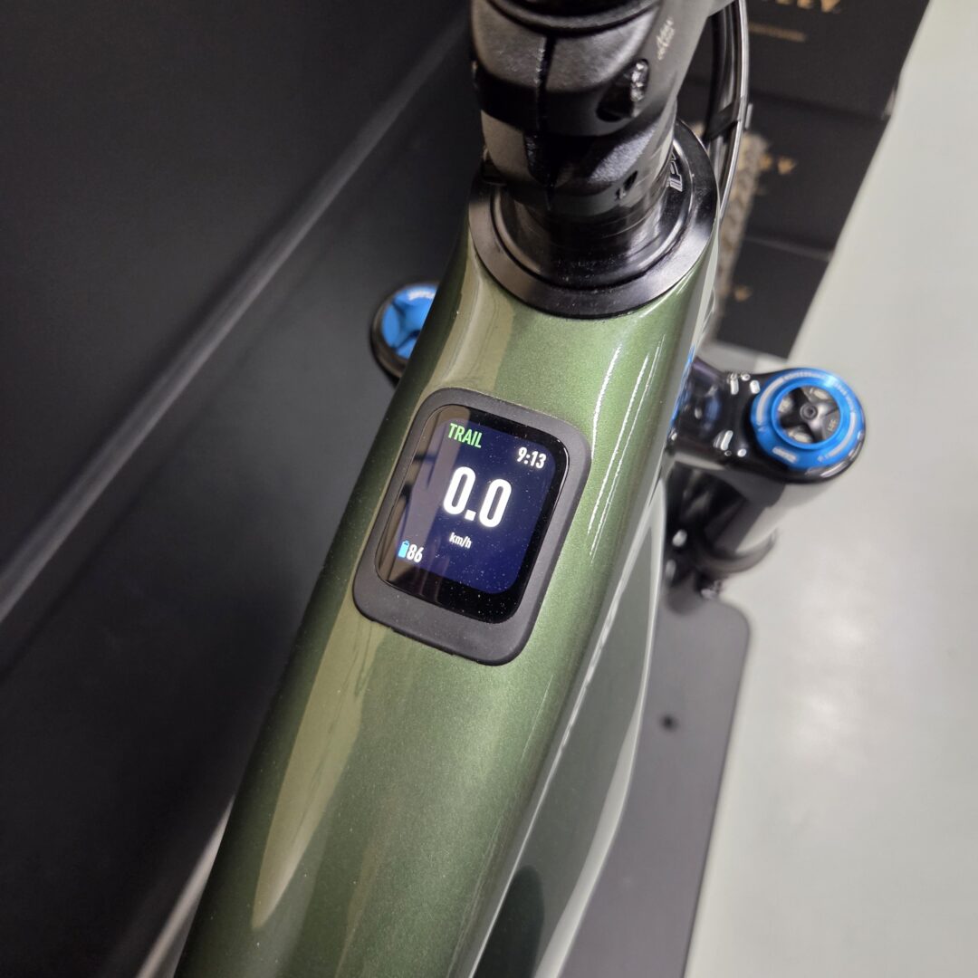Close-up of a green bicycle frame and handlebars with a digital display showing 'TRAIL' and '0.0 km/h' as the bike is stationary, blue adjustment dial visible nearby.