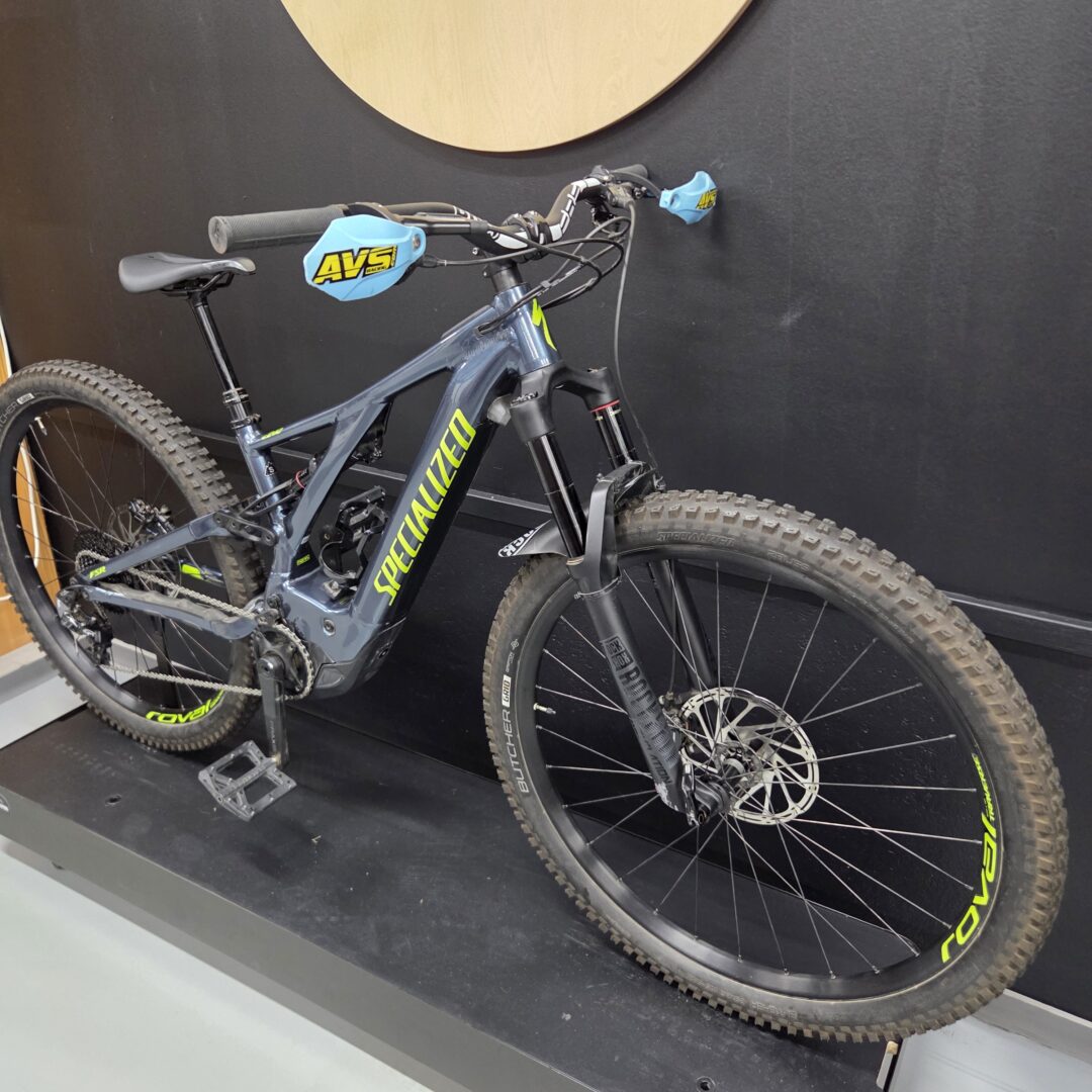 Mountain bike on display with blue AVS-branded handlebar and brake covers, mounted against a dark wall.