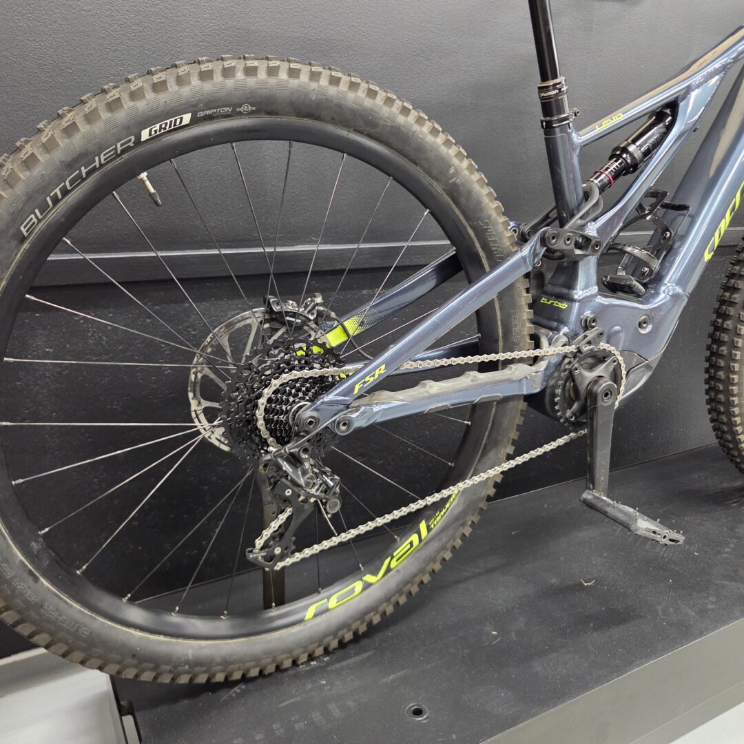 Close-up of a mountain bike's rear wheel and drivetrain, showing the cassette, chain, and frame against a dark wall.