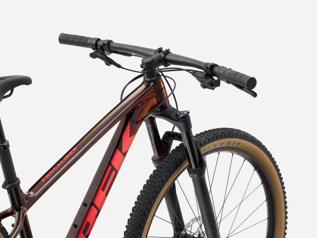 Close-up of a mountain bike's front end: brown frame, black handlebars with grips and brake levers, and a front suspension fork with a knobby tire visible on the right.
