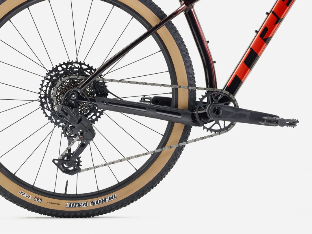 Close-up of a mountain bike's rear wheel showing the derailleur, cassette, chain, and tan-walled tire near an orange frame against a light background.
