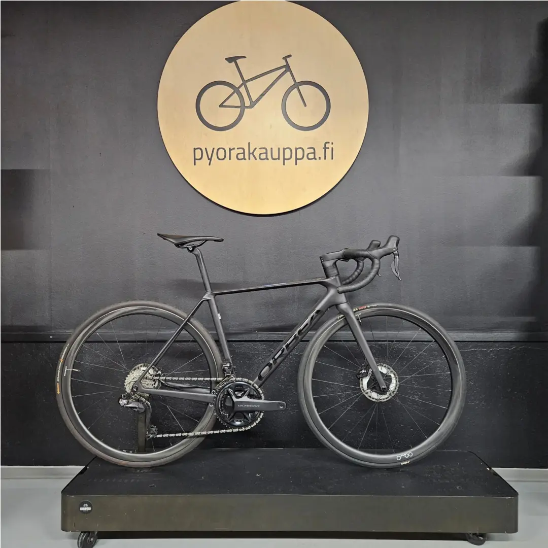 Black road bike on a display platform in a store, with a circular wooden sign reading 'pyorakauppa.fi' on the wall behind it