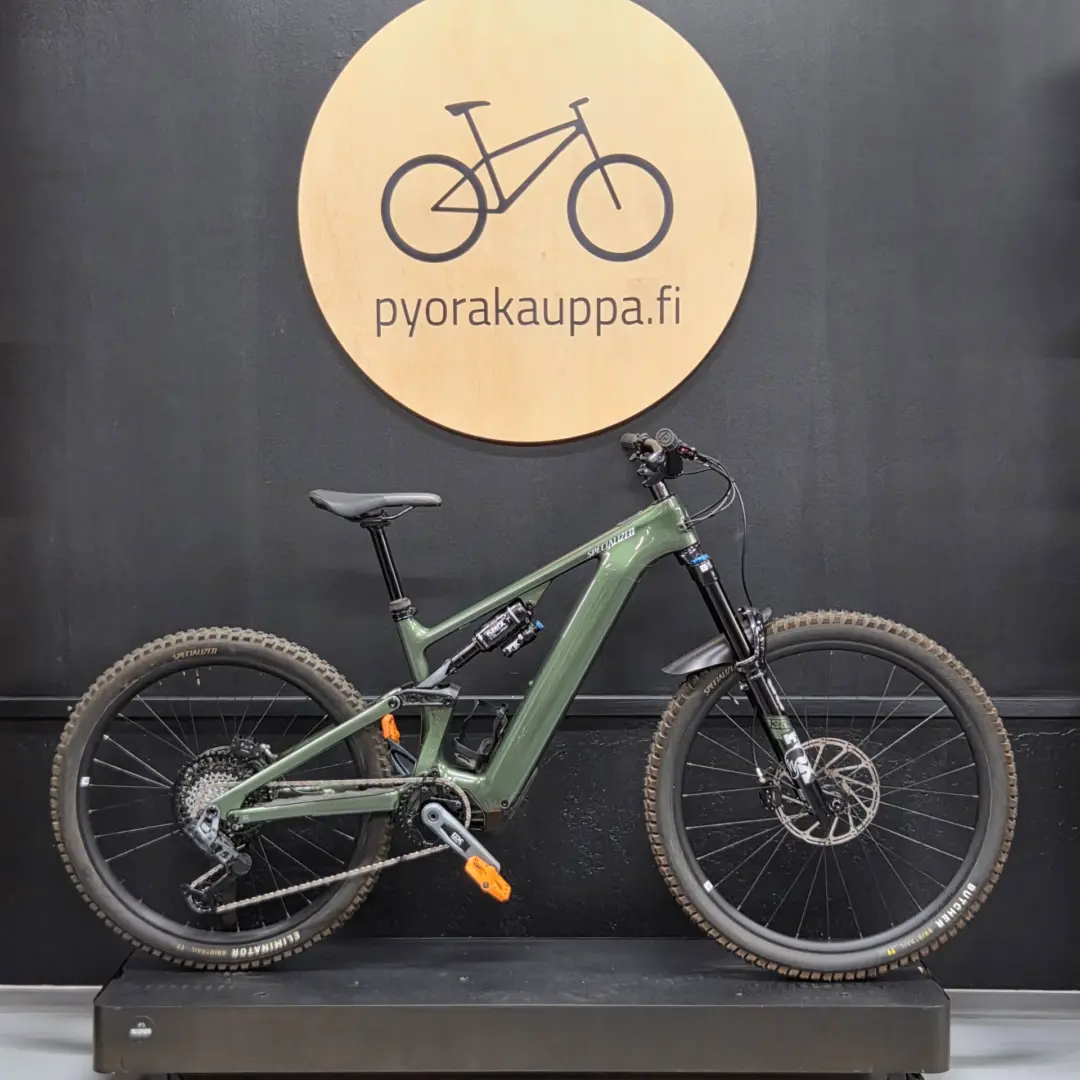 Green mountain bike on a display stand in front of a large circular wooden sign with a bike icon and the text 'pyorakauppa.fi' on a dark wall.