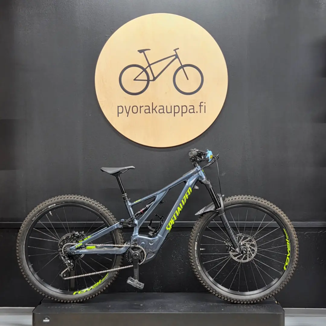 Blue Specialized mountain bike on a display stand against a dark wall, with a round wooden sign reading 'pyorakauppa.fi' behind it.