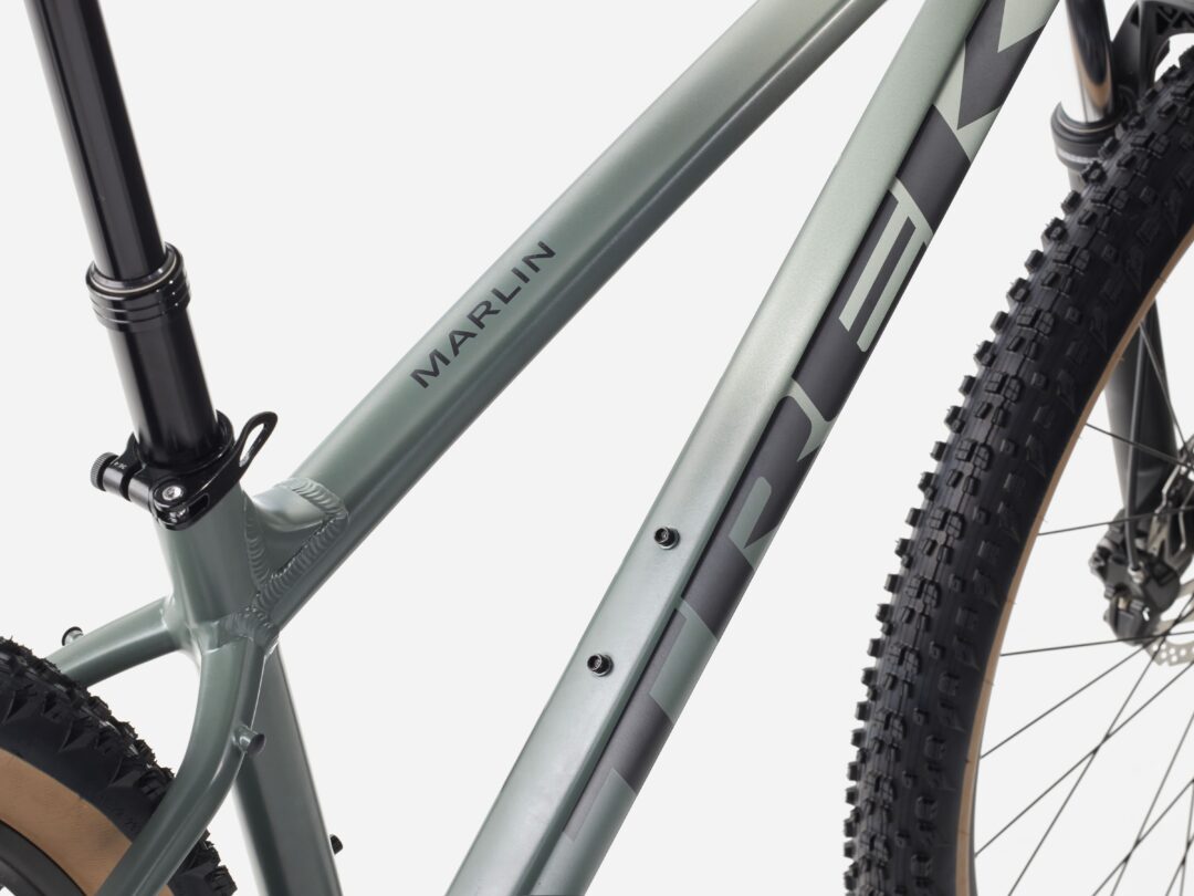 Close-up of a matte green mountain bike frame with the name 'MARLIN', showing part of the rear suspension and a knobby tire in the background.