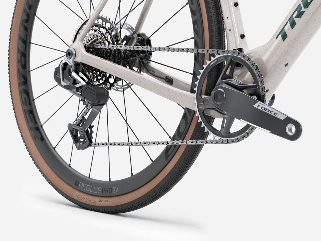 Close-up of a bicycle's rear drivetrain: cassette, derailleur, chain, and crank on a speckled frame with tan tires.