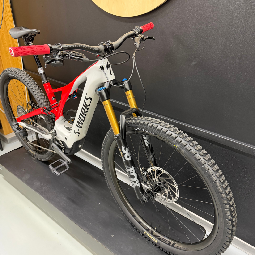 Close-up of a red-and-white full-suspension mountain bike on display, showing red handlebar grips, gold front forks, and knobby tires against a dark wall.