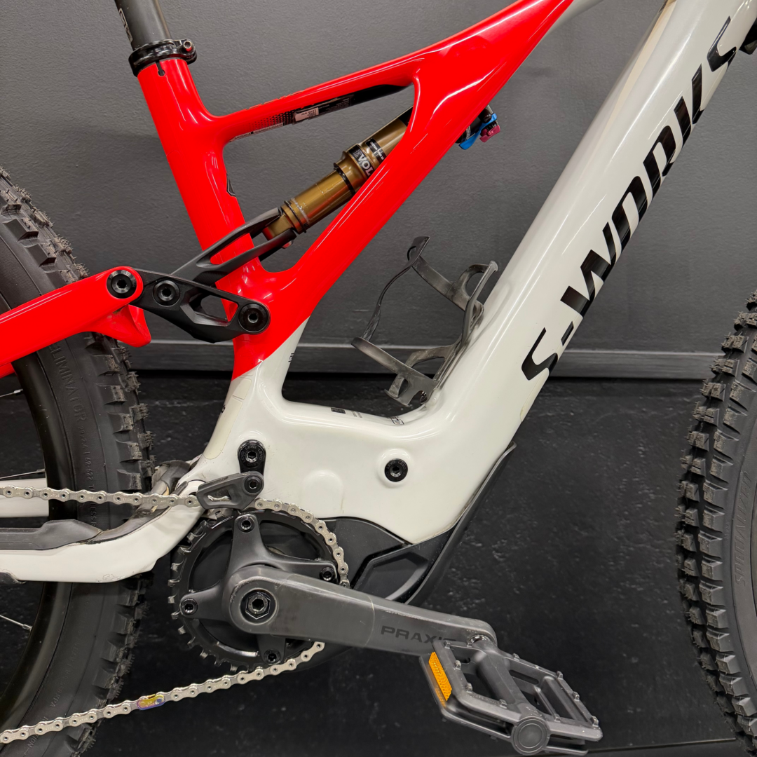 Close-up of a red full-suspension mountain bike frame showing the gold rear shock, drivetrain, and pedals.