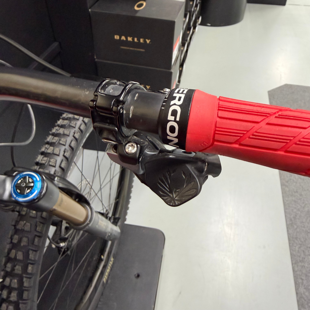 Close-up of a red mountain bike frame and front suspension fork with a knobby tire; in the background is an Oakley-branded black box/drawer unit.