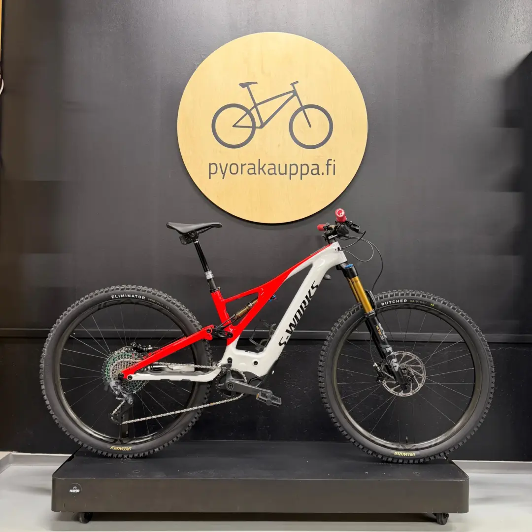 Red and white full-suspension mountain bike on a black display platform against a dark wall, with a round wooden sign showing a bike illustration and 'pyorakauppa.fi'