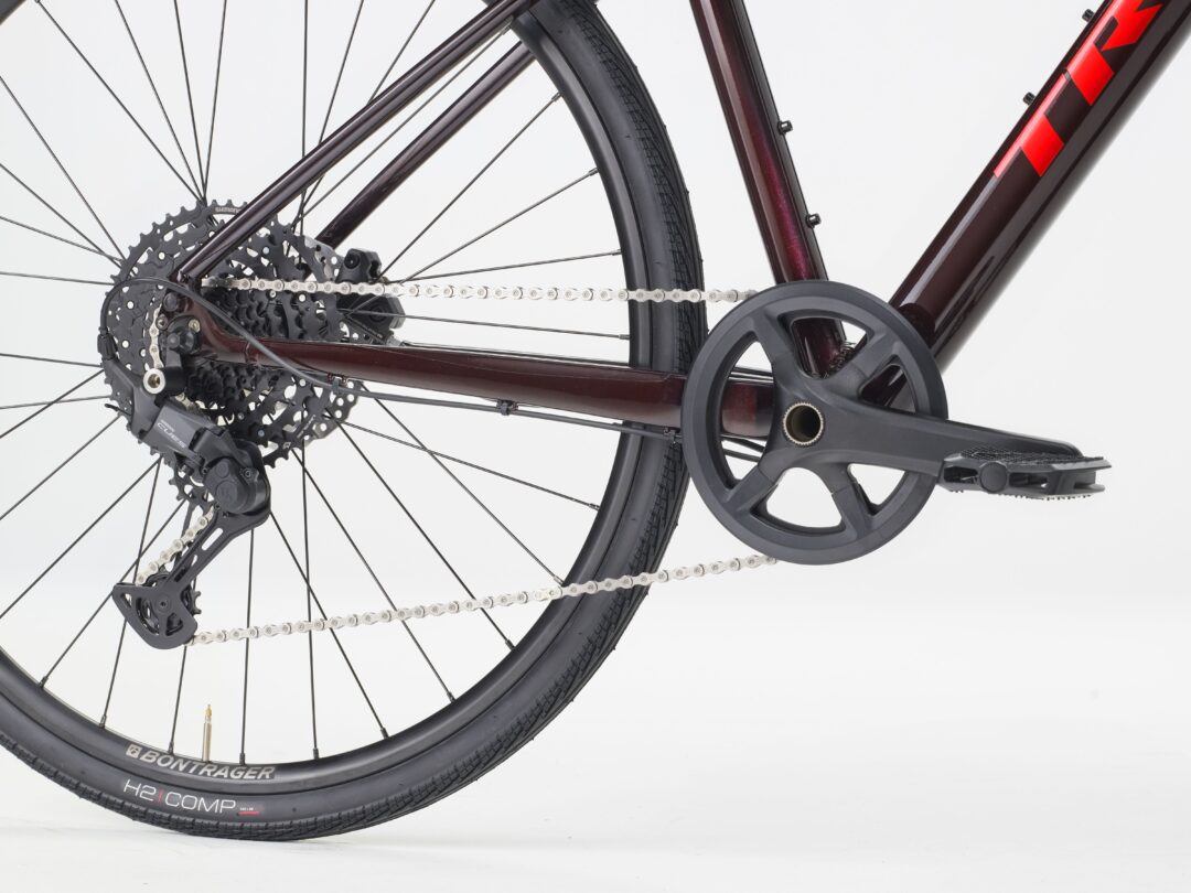 Close-up of a bicycle's rear wheel and drivetrain: cassette, chain, derailleur, and frame tubing visible against a light background.