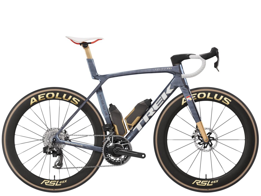 Side view of a high-performance road bicycle with an aero carbon frame, disc brakes, and deep-section wheels labeled 'RSL 65X'; white bar tape and white saddle visible, drivetrain and chainring shown mid-frame.